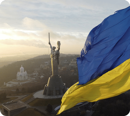 Ukrainian flag waving with Motherland Monument statue and Kyiv cityscape at sunset in background
